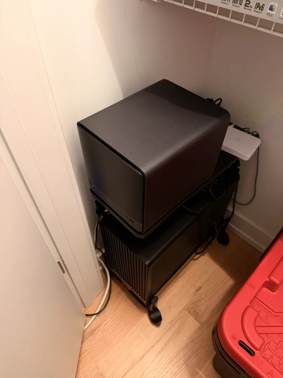 A black cube-shaped NAS sitting on top of a black PC case on casters, in a closet, with a small white network switch behind them