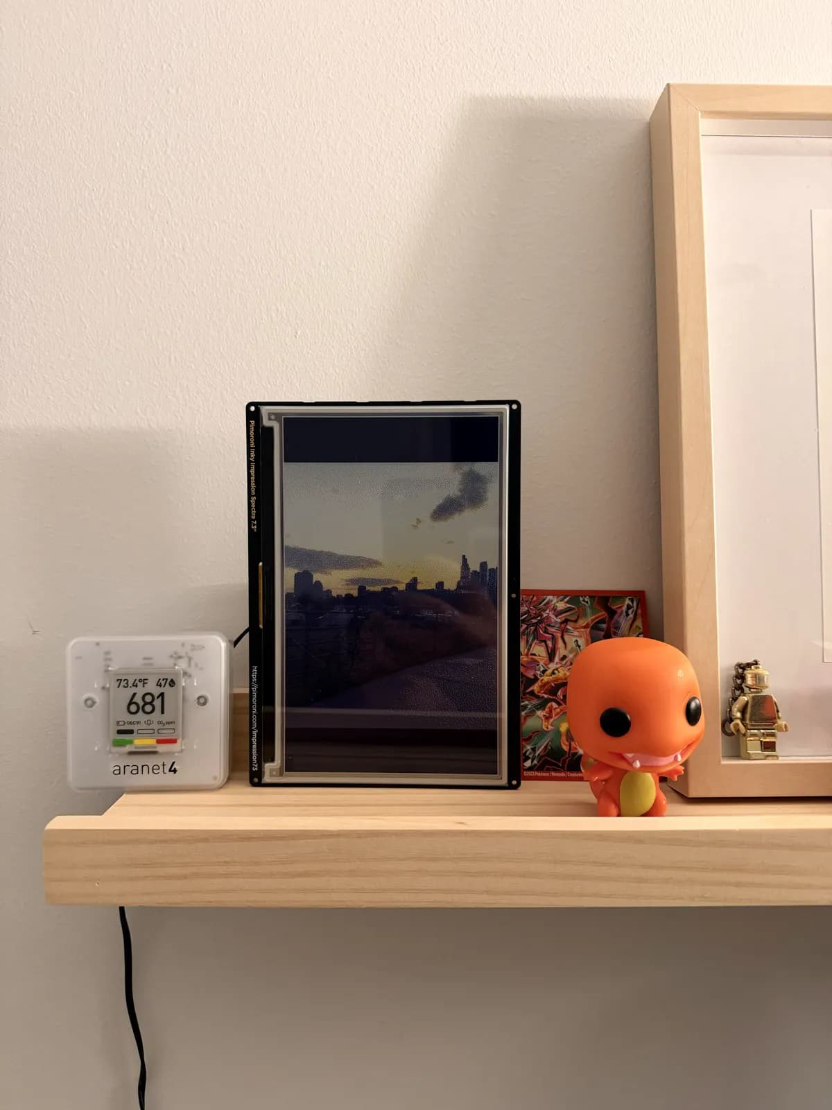 An e-ink picture frame showing a city skyline at sunset on a shelf, next to an Aranet4 sensor reading 73.4°F and a Charmander figure