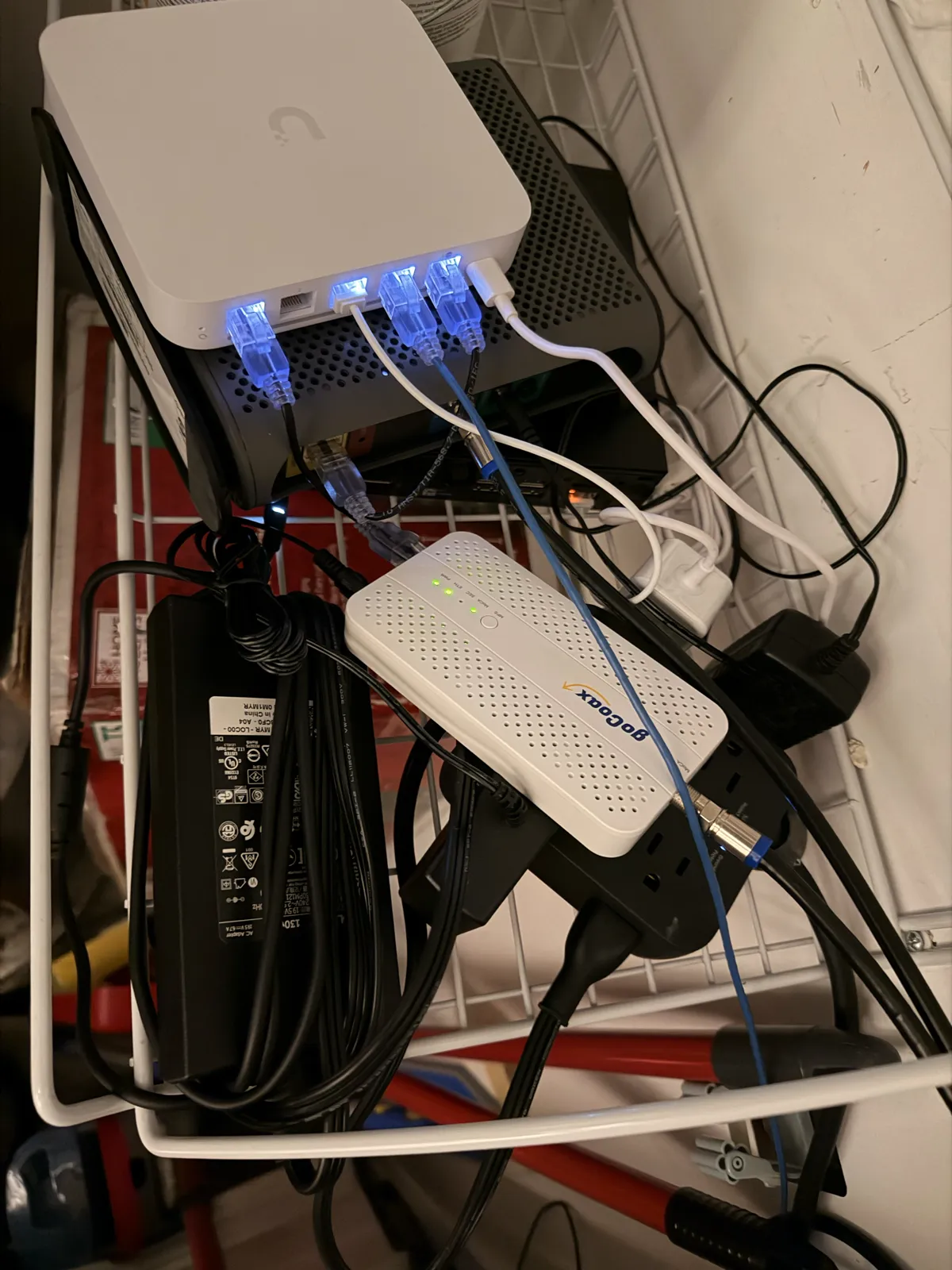 A white UniFi gateway stacked on a black cable modem in a wire basket, with a white goCoax adapter and a power strip below